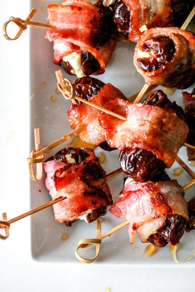 This is an overhead image of bacon wrapped dates skewered on a toothpick. The dates lay on a white rectangle plate on a white counter.