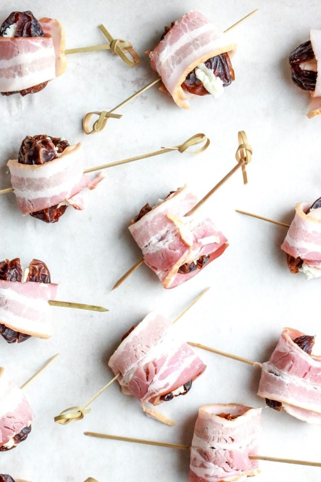 This is an overhead image of dates stuffed with goat cheese and wrapped in bacon. A toothpick skewers through each bacon wrapped date. The dates lay on a white piece of parchment paper.