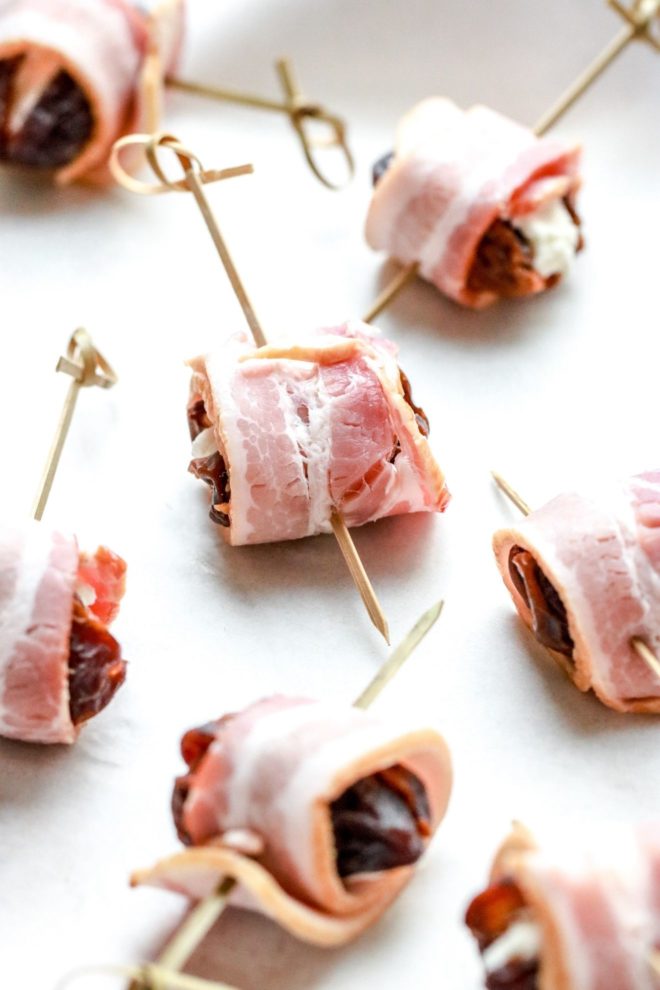 This is a side view of a date stuffed with cheese and wrapped in bacon. A toothpick skewers through the date. The dates sit on a white piece of parchment paper.
