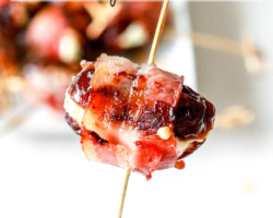 This image shows a hand holding a toothpick that has a bacon wrapped date on it. The date is stuffed with cheese and a plate of more dates is blurred in the background. Text overlay reads "bacon wrapped dates with goat cheese."