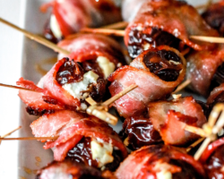 This is a side view of a white rectangle plate with a bunch of bacon wrapped dates on it. The dates are skewered with toothpicks and the plate sits on a white counter. Text overlay reads "bacon wrapped dates with goat cheese."