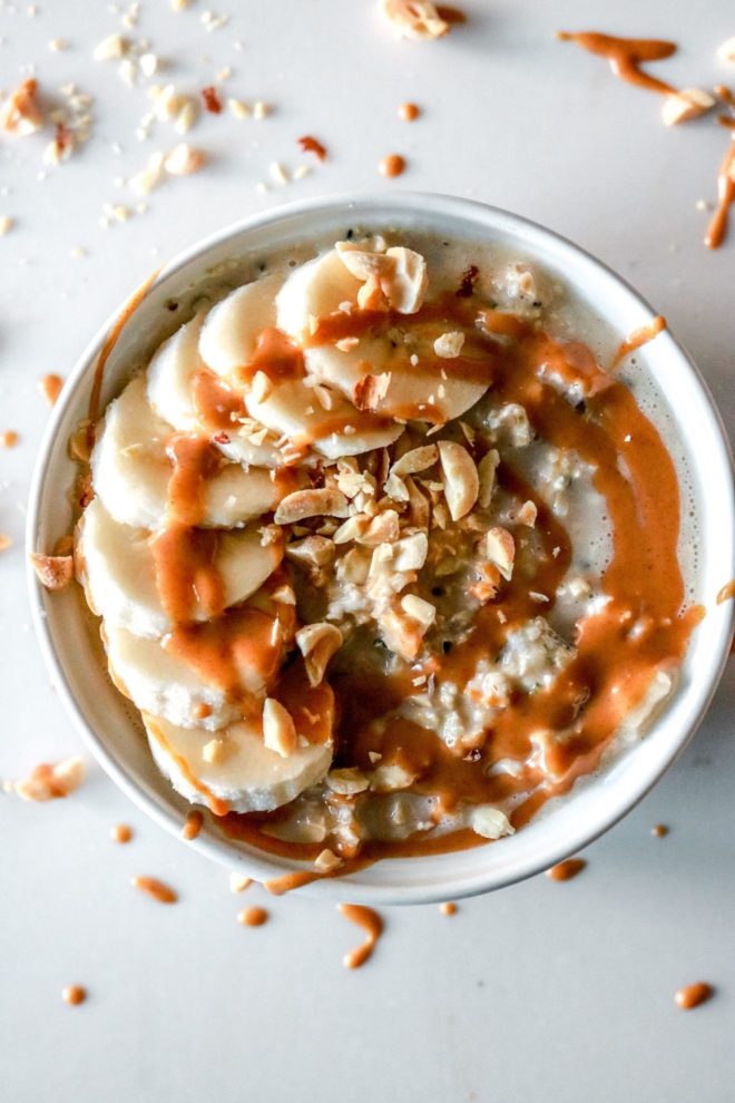 This is an overhead image of a bowl of oats. The oats are topped with sliced banana, a drizzle of peanut butter, and chopped peanuts. The bowl sits on a white counter.