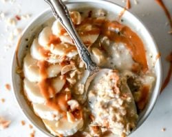 This is an overhead image of a white bowl filled with oatmeal. The oatmeal is topped with sliced banana, a drizzle of peanut butter, and chopped peanuts. A spoonful of oats lays across the top of the bowl.