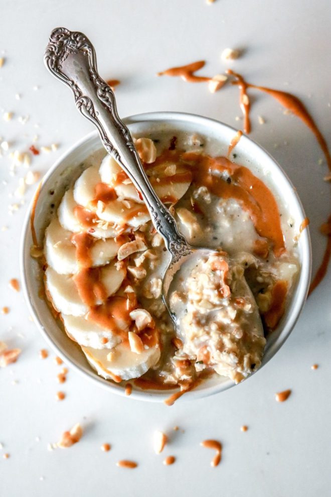 This is an overhead image of a white bowl filled with oatmeal. The oatmeal is topped with sliced banana, a drizzle of peanut butter, and chopped peanuts. A spoonful of oats lays across the top of the bowl.