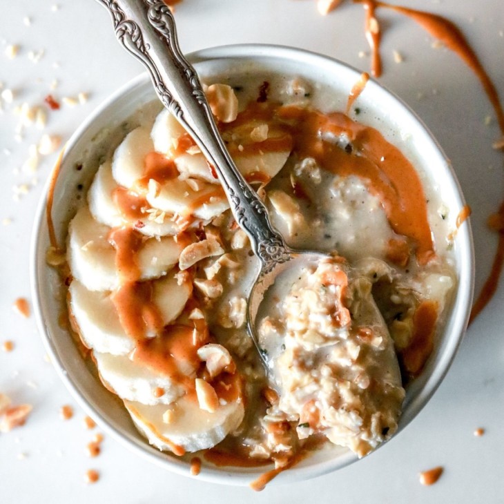 This is an overhead image of a white bowl filled with oatmeal. The oatmeal is topped with sliced banana, a drizzle of peanut butter, and chopped peanuts. A spoonful of oats lays across the top of the bowl.