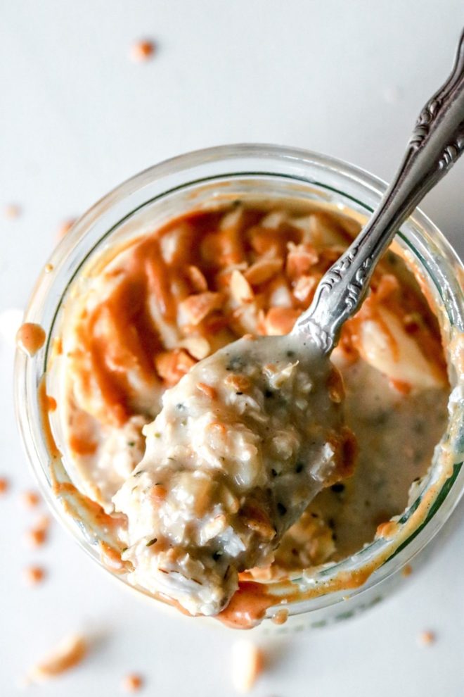 This is an overhead image of a jar of oatmeal. The jar sits on a white counter. The oatmeal is topped with bananas and peanut butter. A spoon full of oatmeal lays across the top of the jar.
