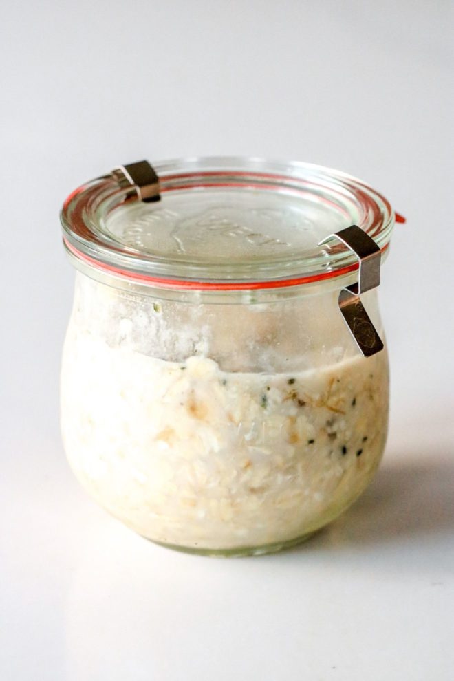 This image is a side view of a weck jar with oatmeal inside. The jar has a lid on it and sits on a white counter.