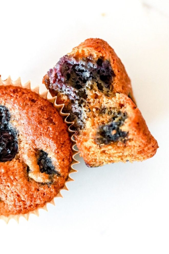 This is an overhead image of a blueberry muffin with a bite taken out of it. The muffin lays on it side, leaning up against another muffin on a white counter.