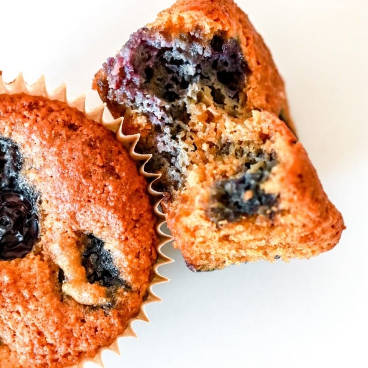 This is an overhead image of a blueberry muffin with a bite taken out of it. The muffin lays on it side, leaning up against another muffin on a white counter.