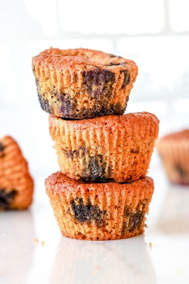 This is a side view of a stack of three blueberry muffins sitting on a white counter with other blueberry muffins blurred in the background.