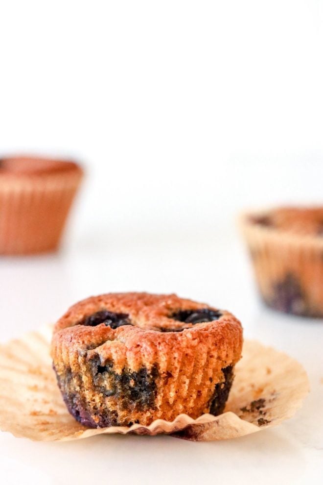 This is a side view of a blueberry muffin with the wrapper pulled off. The muffin sits on a white counter with other muffins blurred in the background.