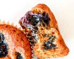 This is an overhead image of a blueberry muffin with a bite taken out of it. The muffin lays on it side, leaning up against another muffin on a white counter. Text overlay reads "gluten free blueberry muffins."