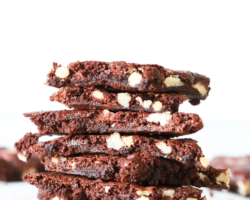 This image is a side view of a stack of brownie brittle with nuts in it. The stack sits on a white counter with other brownie brittle pieces blurred in the background. Text overlay reads "homemade brownie brittle".