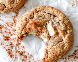 This image is an overhead view of a cookie with a bite taken out of it. The cookie lays on a piece of parchment paper and has chunks of white chocolate and a sprinkle of salt.
