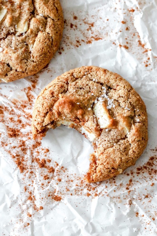 This image is an overhead view of a cookie with a bite taken out of it. The cookie lays on a piece of parchment paper and has chunks of white chocolate and a sprinkle of salt.