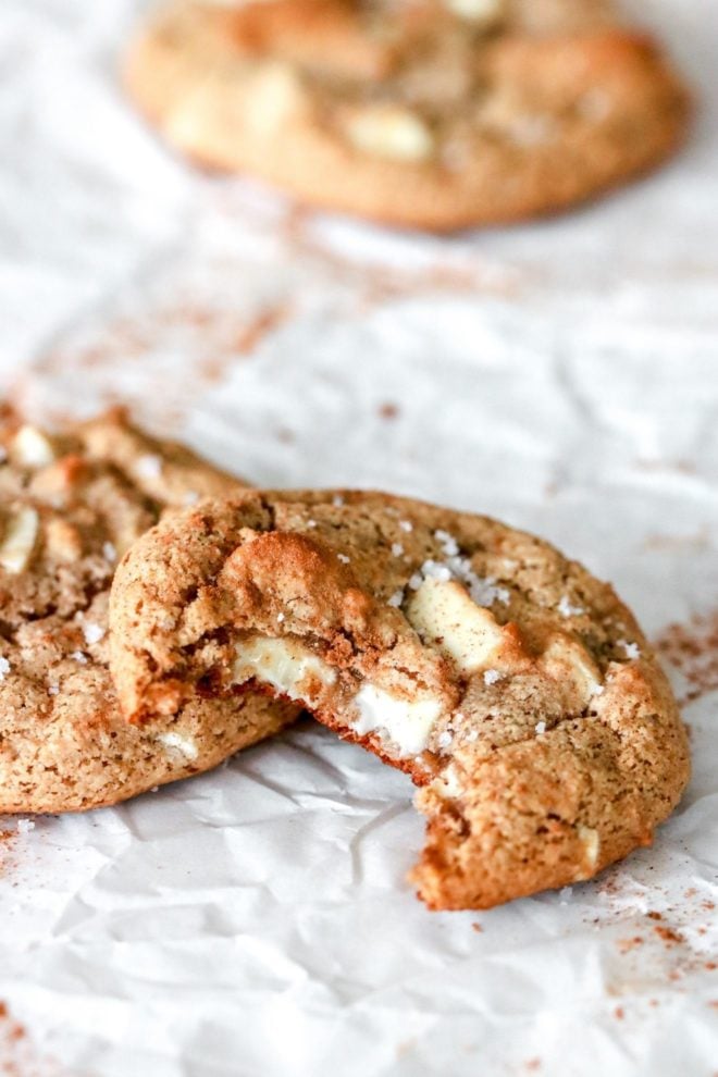 This is a side view of a cookie with a bite taken out of it. The cookie has white chocolate chunks and salt sprinkled on top. It leans against another cookie and another cookie is blurred in the background.