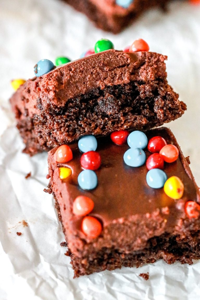 This is an image of a cosmic brownie with a layer of fudgey brownie, a layer of chocolate ganache frosting, and colorful chocolates on top. The brownie is leaning against another brownie and both sit on a piece of white parchment paper.