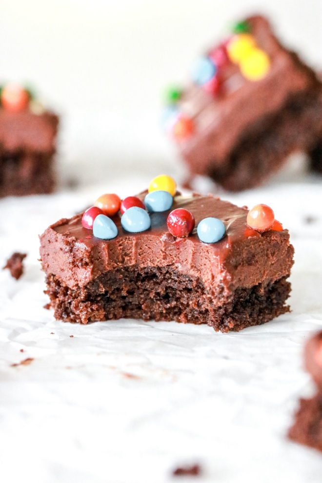 This image is a side view of a cosmic brownie with a bite taken out of it. The brownie has a brownie base layer, with a thick layer or chocolate frosting, and is topped with colorful candies. The brownie sits on a white counter with other brownies blurred in the background.