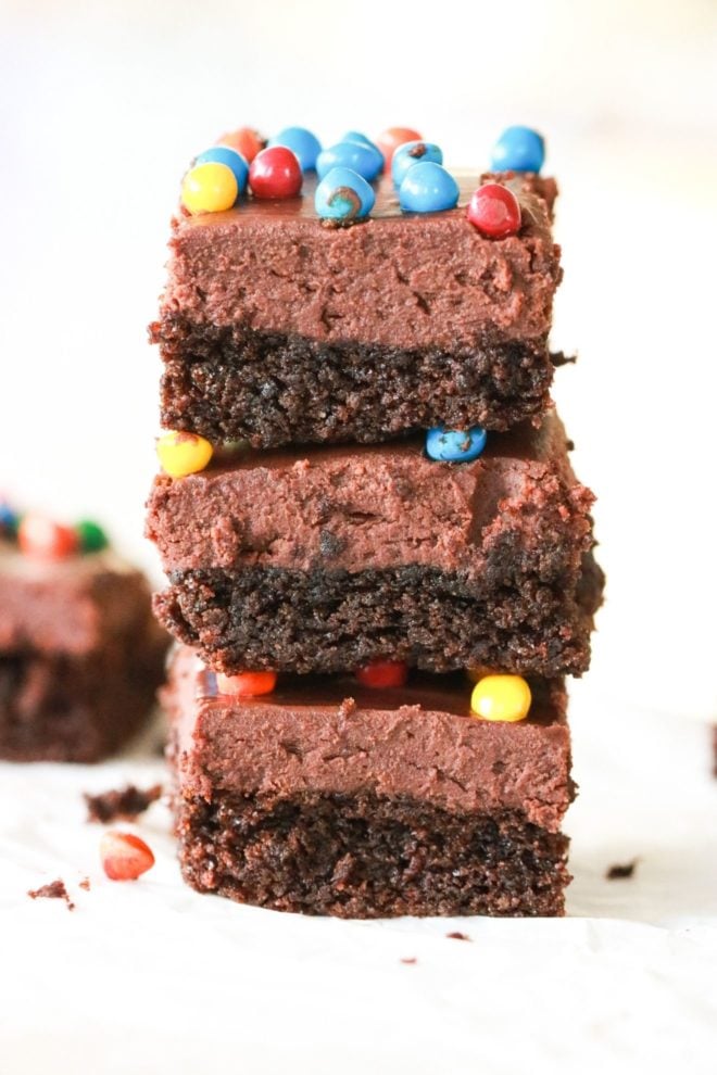 This image is a side view of a stack of three cosmic brownies. The brownies have a base layer of brownie, a layer of chocolate frosting, and a top layer of colorful candies. The brownies sit on a piece of white parchment paper and other brownies are blurred in the white background.
