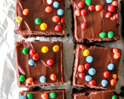 This is an overhead image of cosmic brownies with chocolate icing and colorful candies sprinkled on top. The brownies lay on a white piece of parchment paper. text overlay reads "fudgey cosmic brownies."