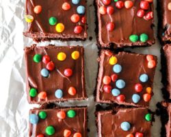 This is an overhead image of brownies with chocolate frosting and colorful candies on top. The brownies sit on a white piece of parchment paper.