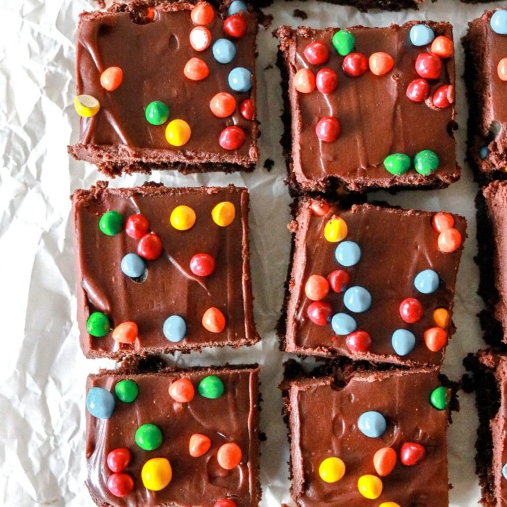This is an overhead image of brownies with chocolate frosting and colorful candies on top. The brownies sit on a white piece of parchment paper.