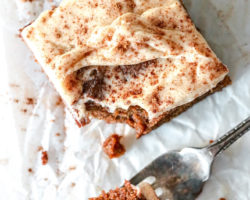 overhead image of a square slice of gingerbread cake with vanilla icing and a sprinkle of cinnamon. a fork takes a bite out of the cake slice and lays on the wrinkled white piece of parchment paper.