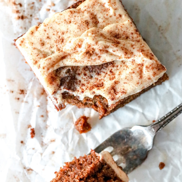overhead image of a square slice of gingerbread cake with vanilla icing and a sprinkle of cinnamon. a fork takes a bite out of the cake slice and lays on the wrinkled white piece of parchment paper.