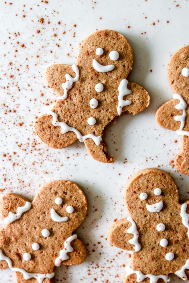 This is an overhead image of a batch of gingerbread men cookies with decorative icing. The cookies lay on a white background with a sprinkle of cinnamon.