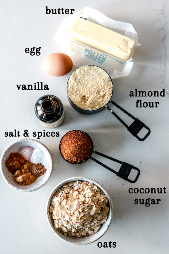 overhead image of butter, an egg, a bottle o vanilla extract, almond flour, coconut sugar, salt and spices, and oats laid out on a white counter