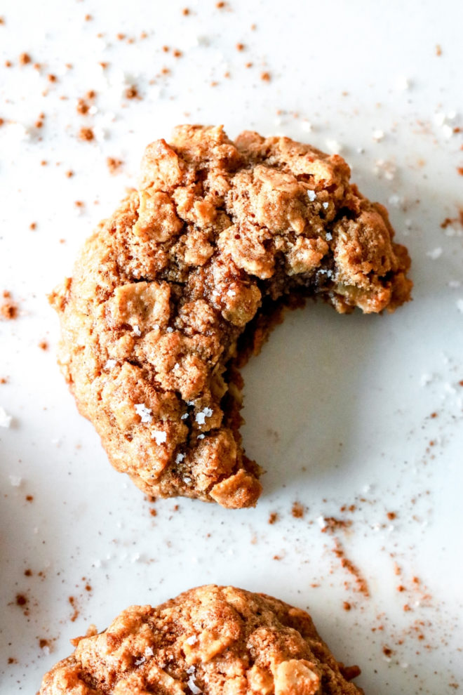 overhead image of an oatmeal cookie with a bite taken out laying on a white counter and ground cinnamon sprinkled around it.