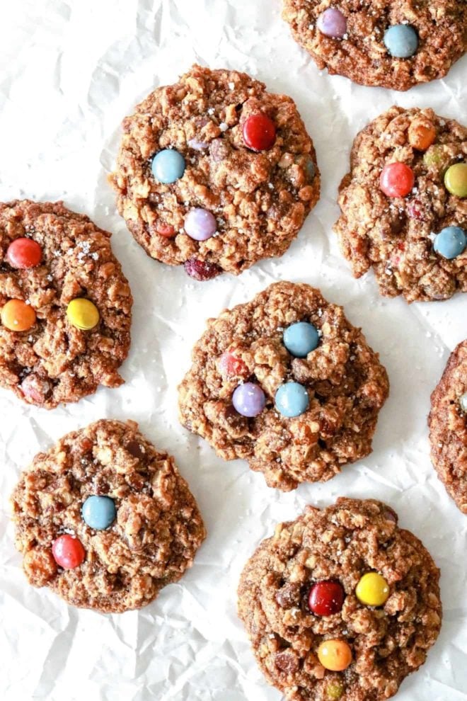 This is an overhead image of a bunch of monster cookies with oats and M&Ms. The cookies lay on a white piece of parchment paper and are sprinkled with salt.
