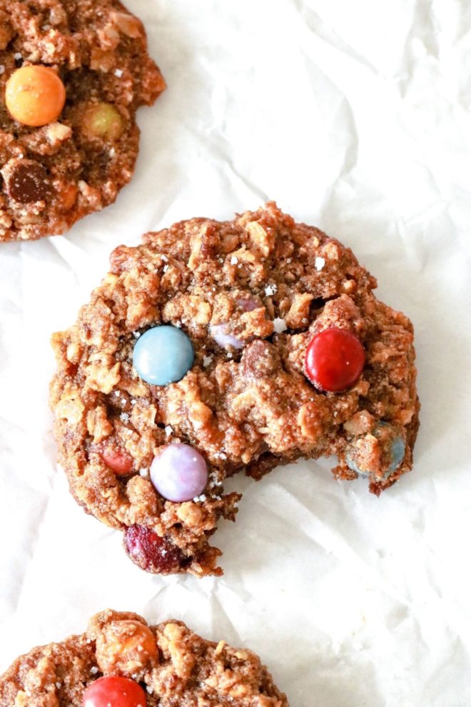 This is an overhead image of a monster cookie with M&Ms and oats with a bite taken out. The cookie lays on a piece of parchment paper.