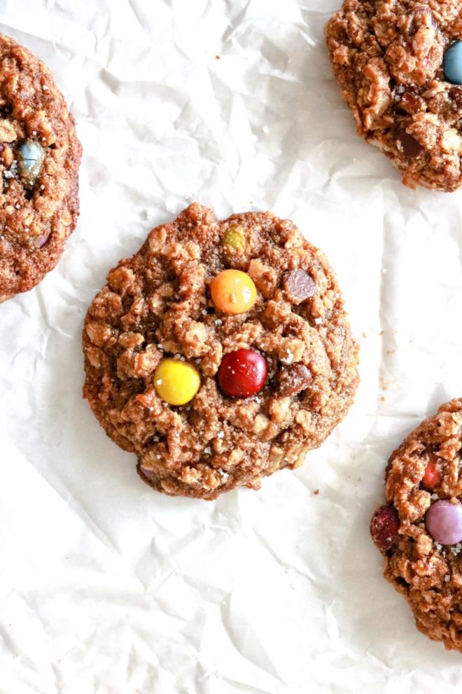 This is an overhead image of monster cookies laying on a white piece of parchment paper. The cookies have oats and M&Ms visible and are sprinkled with some salt.