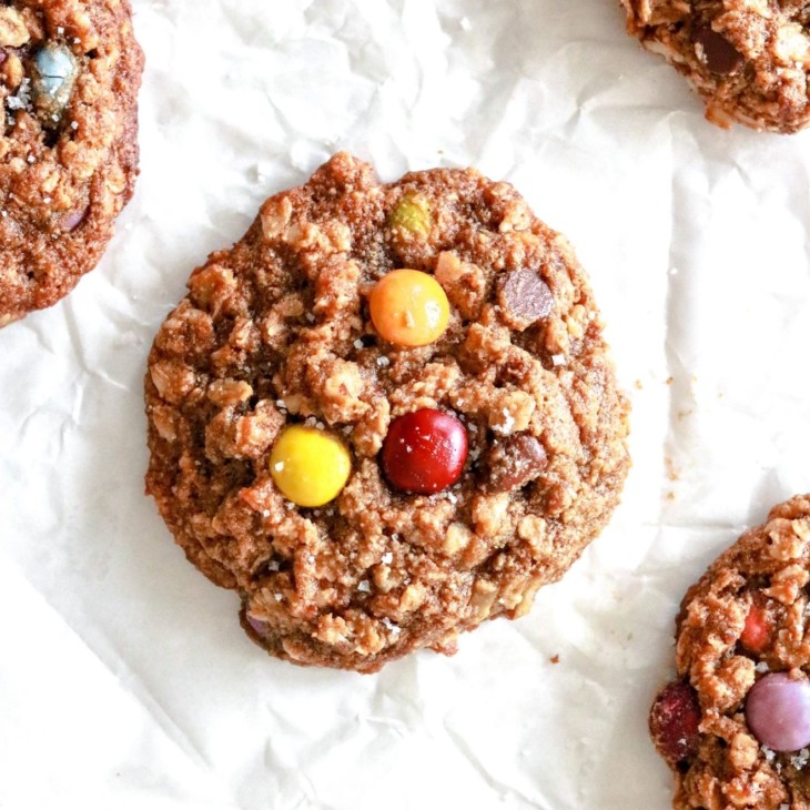 This is an overhead image of gluten free monster cookies laying on a white piece of parchment paper. The cookies have oats and M&Ms visible and are sprinkled with some salt.