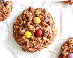 This is an overhead image of monster cookies laying on a white piece of parchment paper. The cookies have oats and M&Ms visible and are sprinkled with some salt. Text overlay reads "healthy monster cookies."