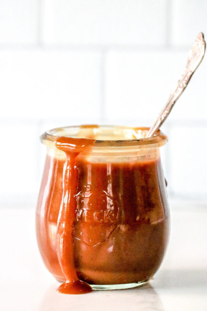 side view of a small glass jar with caramel inside and dripping off the edge. a spoon sticks out of the jar and the jar sits on a white counter with a white background.