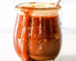 side view of a small glass jar with caramel inside and dripping off the edge. a spoon sticks out of the jar and the jar sits on a white counter with a white background.