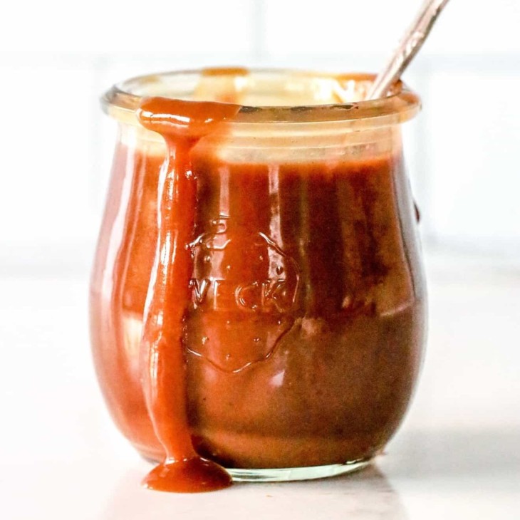 side view of a small glass jar with caramel inside and dripping off the edge. a spoon sticks out of the jar and the jar sits on a white counter with a white background.