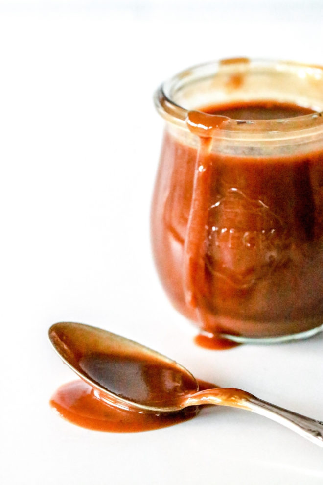 side view of a spoon with caramel sauce spilling onto the white counter with a small glass jar of caramel next to it.