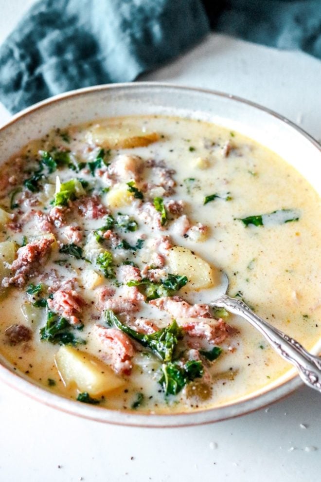 This is a side image of a soup bowl with a cream broth, sausage, kale, and potatoes. A spoon scoops a potato and leans against the side of the bowl. The bowl sits on a white counter with a blue napkin blurred in the background.