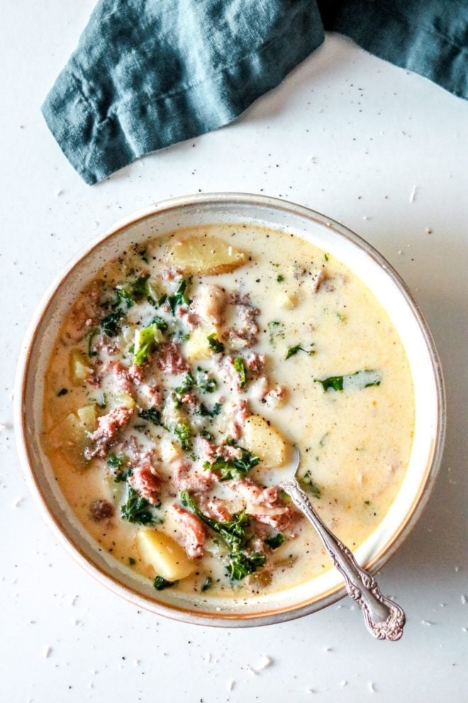 This is an overhead image of a soup bowl on a white counter with a blue dish towel at the top of the image. The soup bowl has a cream broth with meat, potatoes, and kale inside.