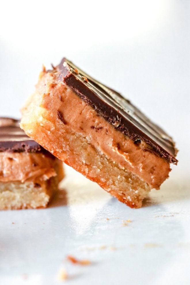 side view of a peanut butter millionaire bar leaning against another bar. both bars sit on a white counter and have three layers: a shortbread layer, peanut butter layer, and chocolate layer.