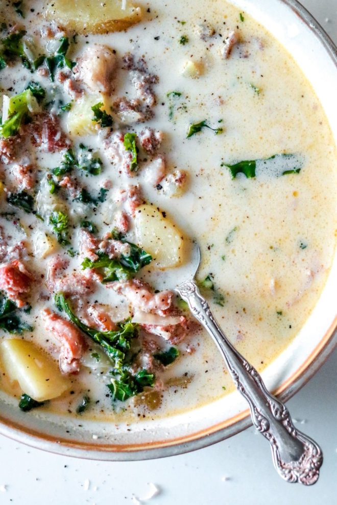 This is a close up image of a spoon leaning against the side of the bowl. The spoon dips down into a creamy broth with sausage, kale, and potatoes. The spoon scoops up a small potato piece.