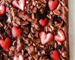 This is an overhead image of brownies on a white piece of parchment paper. The brownie is topped with chopped chocolate, strawberry jam, and sliced strawberries shaped like hearts.