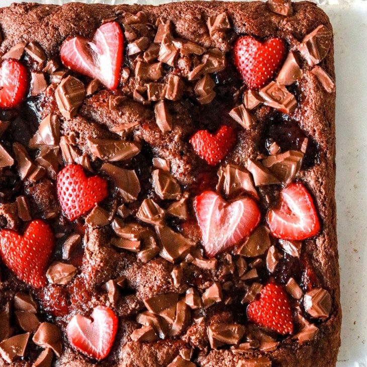 This is an overhead image of brownies on a white piece of parchment paper. The brownie is topped with chopped chocolate, strawberry jam, and sliced strawberries shaped like hearts.