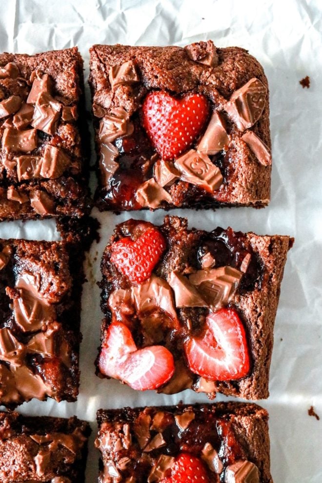 This is an overhead image of brownies cut in squares. The brownies sit on a white piece of parchment paper and are topped with chopped chocolate, strawberry jam, and sliced strawberries.