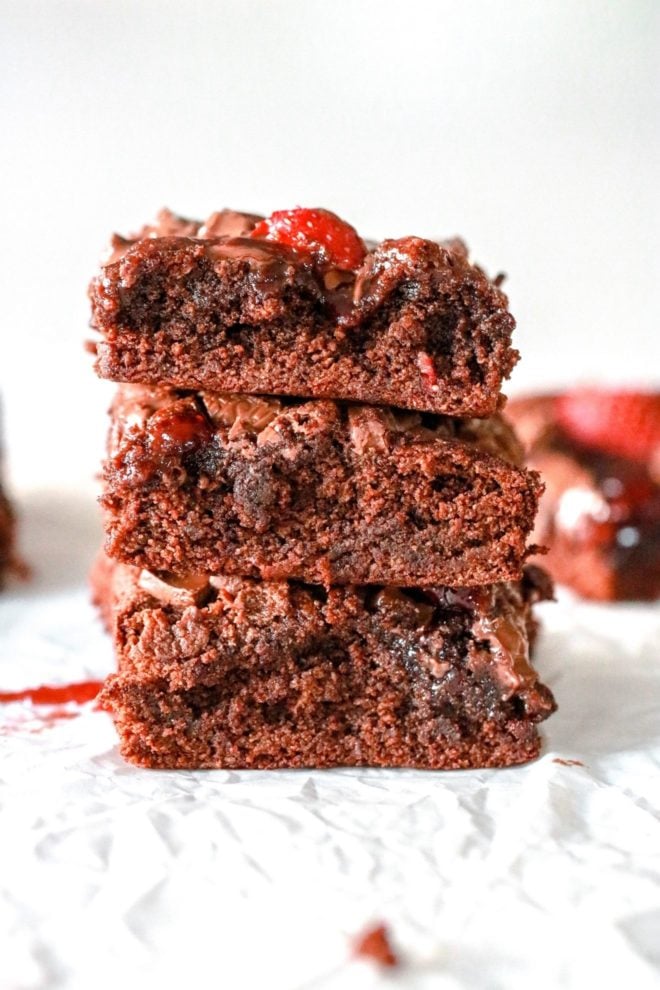 This is a side view of a stack of three brownies. The stack sits on top of a white counter and a white background. More brownies are blurred in the background.
