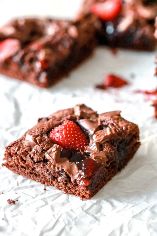 This is a side view of of a brownie topped with jam and a fresh strawberry. The brownie sits on a white piece of parchment paper and more brownies are blurred in the background.