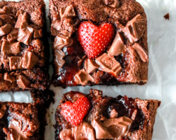 This is an overhead image of cut brownies on a white piece of parchment paper. The brownies have chopped chocolate, jam, and heart shaped strawberries on top. Text overlay reads "adorable & fudgey strawberry brownies"